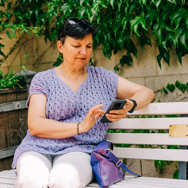Žena sedi na klupi u parku, nosi ljubičastu majicu i svetle pantalone, dok gleda u svoj mobilni telefon. U pozadini su biljke i delimično zid.