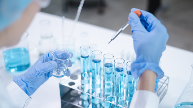 Osoba u laboratorijskoj odori i plavim rukavicama drži čašu s tečnošću dok pipetom dodaje kapljice u epruvete sa plavom tečnošću. U pozadini se nalaze razne laboratorijske posude.
