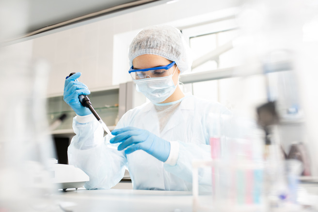 Laborant u zaštitnoj opremi, uključujući masku, rukavice i naočare, koristi pipetu za merenje tečnosti u laboratoriji. U pozadini se vide laboratorijski instrumenti i stakleni sudovi.