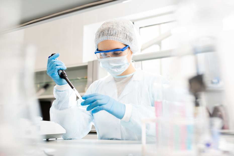 Laborant u zaštitnoj opremi, uključujući masku, rukavice i naočare, koristi pipetu za merenje tečnosti u laboratoriji. U pozadini se vide laboratorijski instrumenti i stakleni sudovi.
