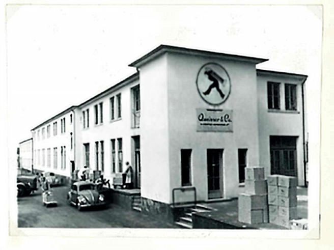 Zgrada kompanije sa natpisom "Quinvar & Co." na fasadi. Vidi se nekoliko ljudi ispred zgrade, kao i automobili parkirani u blizini. U okolini su kutije, što sugeriše poslovnu aktivnost. Fotografija je crno-bela.
