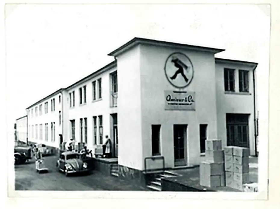 Zgrada kompanije sa natpisom "Quinvar & Co." na fasadi. Vidi se nekoliko ljudi ispred zgrade, kao i automobili parkirani u blizini. U okolini su kutije, što sugeriše poslovnu aktivnost. Fotografija je crno-bela.