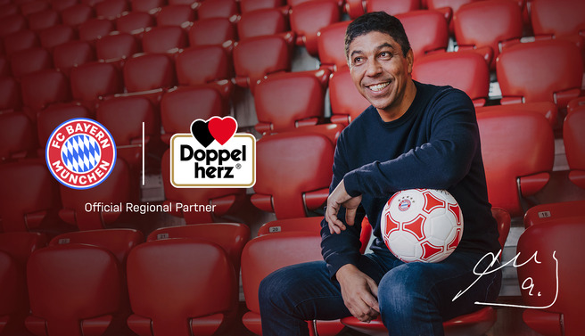 Osoba sedi na stadionu sa crvenim sedištima, drži fudbalsku loptu. U pozadini su logotipi fudbalskog kluba FC Bayern München i brenda Doppelherz. Prikazuje se i natpis "Official Regional Partner".