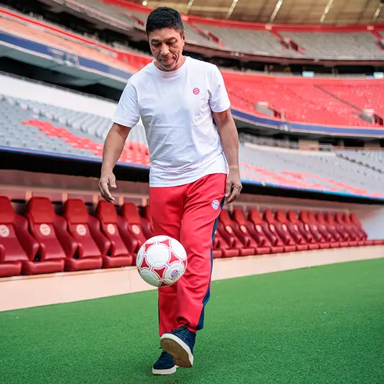 Na slici je muškarac u beloj majici i crvenim trenerkama, kako drži fudbalsku loptu na terenu u fudbalskom stadionu. U pozadini se vide prazna mesta za sedenje. Atmosfera je sportska i opuštena.