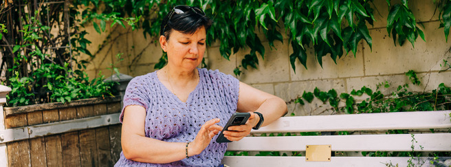 Žena sedi na klupi u dvorištu, obučena u svetlu, džemper s uzorkom, i gleda u svoj mobilni telefon. U pozadini su zelene biljke koje se penju uz zid.