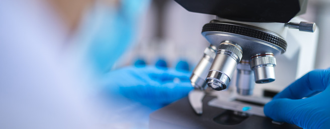Mikroskop sa nekoliko sočiva, na stolu u laboratoriji. Plave rukavice se koriste za podešavanje mikroskopa, dok je u pozadini zamagljena figura nekoga ko radi u laboratoriji.