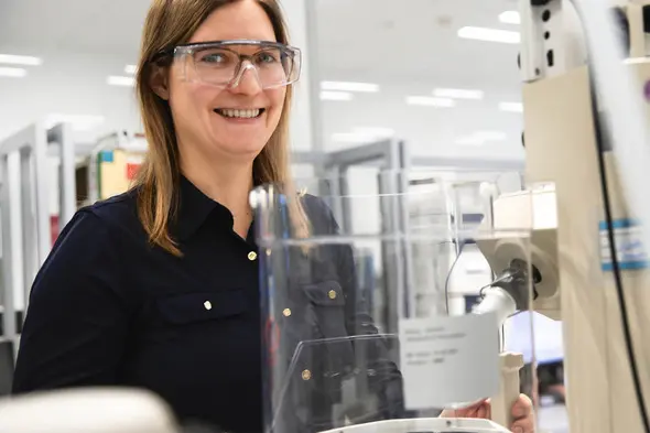 Žena sa smeškom, obučena u tamnu košulju, stoji pored laboratorijske opreme sa zaštitnim naočarima. U pozadini se vide laboratorijski stolovi i oprema.