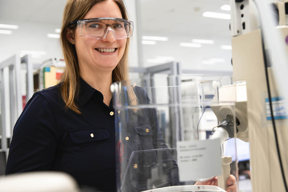 Žena sa smeškom, obučena u tamnu košulju, stoji pored laboratorijske opreme sa zaštitnim naočarima. U pozadini se vide laboratorijski stolovi i oprema.